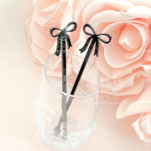 May include: Two black acrylic drink stirrers with bow-shaped tops rest in a clear glass. The stirrers are set against a soft pink background with a large, light pink flower. The stirrers are a decorative addition to cocktails.