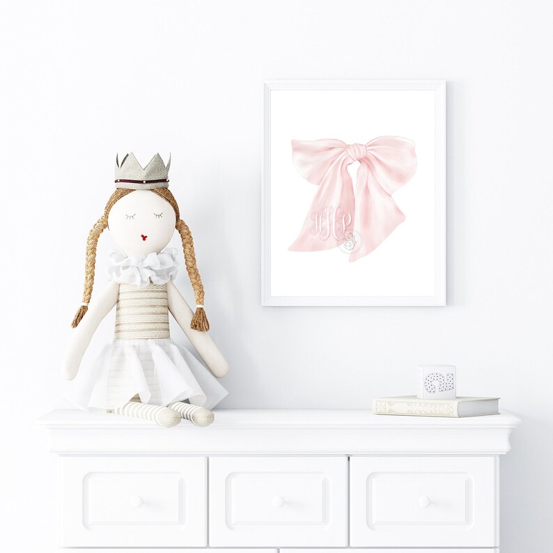 May include: A white dresser with two drawers, a pink bow with a monogram on it in a white frame, and a doll wearing a white dress and a crown.