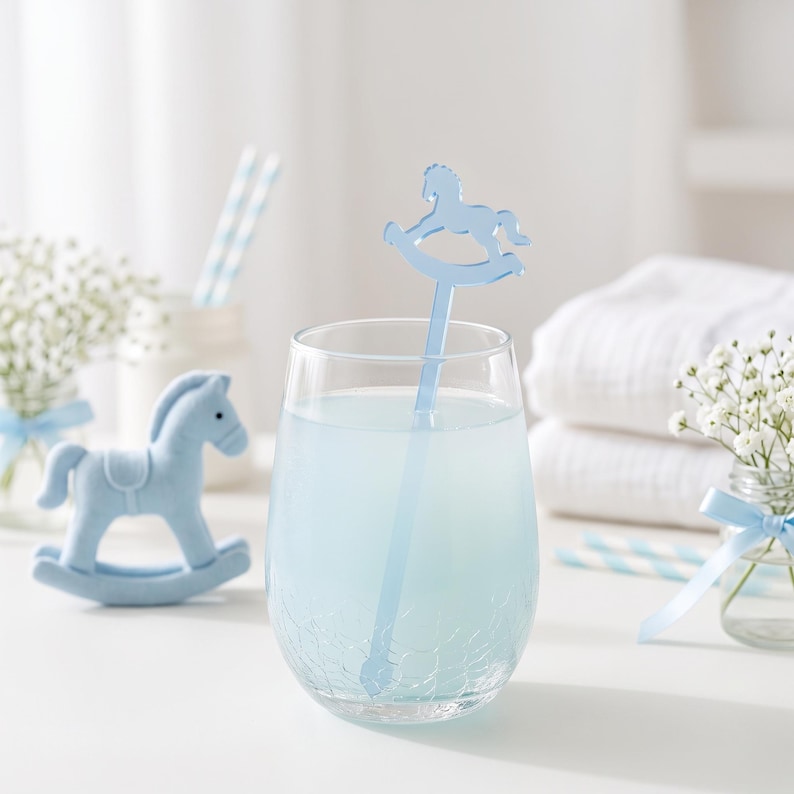 May include: A clear glass tumbler with a crackled texture filled with light blue liquid, featuring a light blue acrylic drink stirrer shaped like a rocking horse. A small, solid light blue rocking horse figurine sits to the left of the glass.
