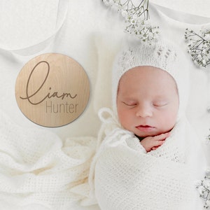 K&ouml;nnte beinhalten: Eine runde Holzplakette mit dem Namen "Liam Hunter", der in kursiver Schrift graviert ist. Ein Neugeborenes schl&auml;ft in einer wei&szlig;en H&auml;keldecke und -m&uuml;tze.