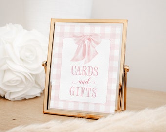 May include: A gold-framed sign with a pink gingham background and a pink bow. The sign reads "Cards and Gifts".