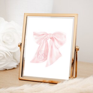 May include: A pink watercolor bow with a white monogram in the center. The bow is in a gold frame.