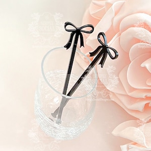 May include: Two black acrylic drink stirrers with bow-shaped tops rest in a clear glass. The stirrers are angled, with one partially submerged. The background features a soft pink rose and a light, blurred backdrop. The stirrers are from Simply Social Designs.