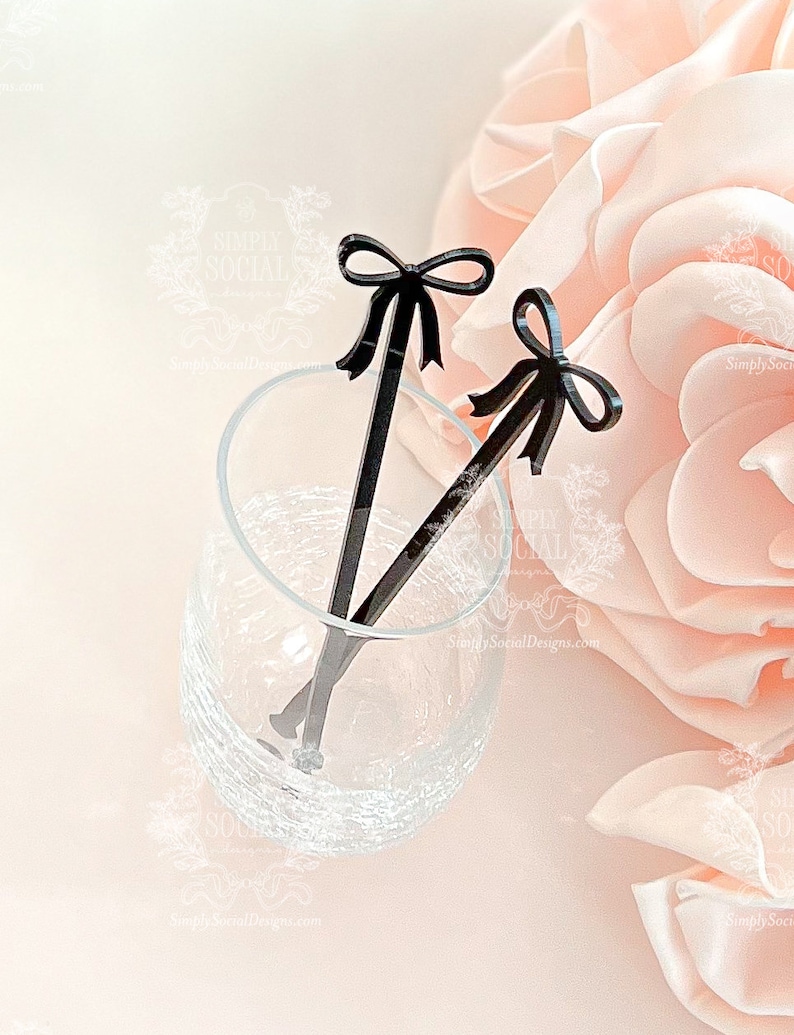 May include: Two black acrylic drink stirrers with bow-shaped tops rest in a clear glass. The stirrers are angled, with one partially submerged. The background features a soft pink rose and a light, blurred backdrop. The stirrers are from Simply Social Designs.