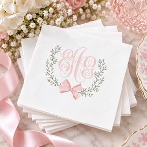 May include: A stack of square white paper napkins featuring a centered design with the letters A and S in a pink cursive monogram. The monogram is framed by a green leafy wreath and a pink ribbon bow at the bottom, printed on the top napkin.