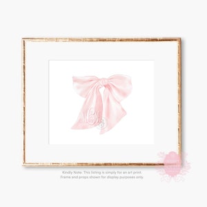 May include: A pink satin bow with a monogram in the center. The bow is on a white background and is framed in a gold frame.