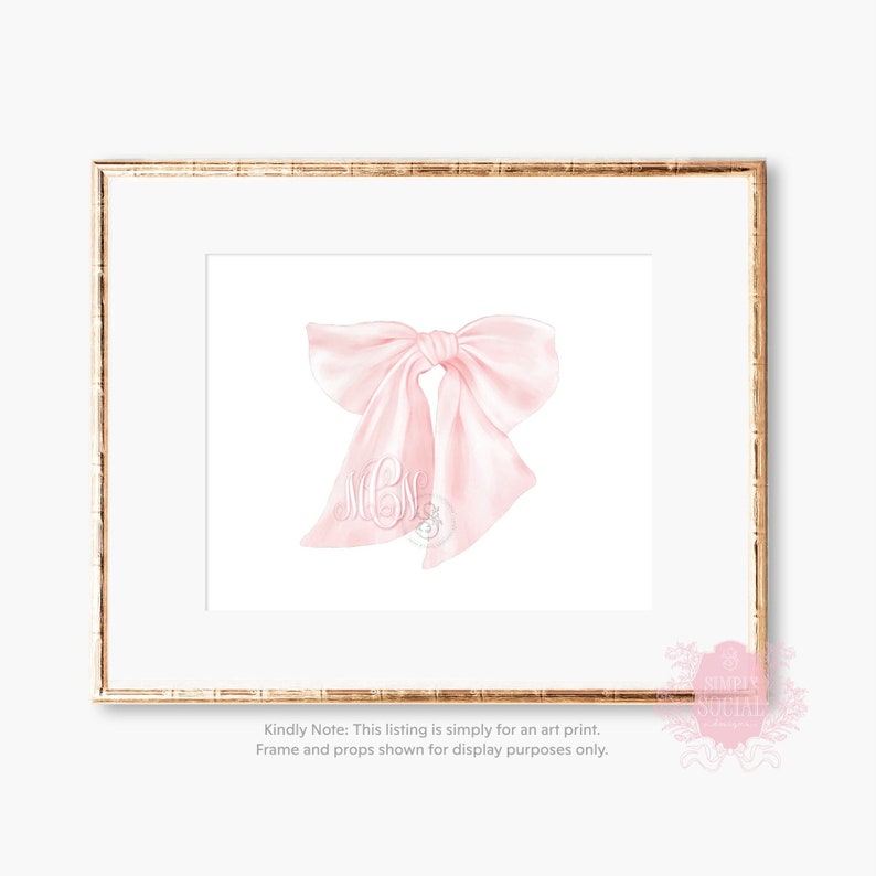 May include: A pink satin bow with a monogram in the center. The bow is on a white background and is framed in a gold frame.