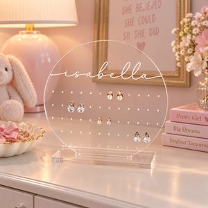 May include: A circular clear acrylic earring holder stand featuring the name Isabella in white script lettering. The stand has a rectangular base and multiple pre-drilled holes for organizing jewelry, with several pairs of earrings displayed.
