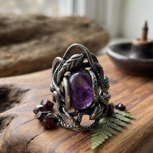 Può includere: Un anello color argento con una pietra ametista viola di forma ovale al centro. La fascia è realizzata con un dettagliato motivo a serpenti intrecciati che avvolge la gemma. Piccole pietre di granato rosso scuro sono posizionate alla base.