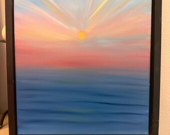 May include: A framed painting depicting a sunrise over a calm blue ocean. The sky features a gradient of soft pink and light blue hues, with bright yellow sun rays radiating upwards from a small, circular yellow sun at the centre of the horizon.