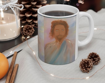 May include: A white ceramic mug featuring a printed artistic illustration of a person with shoulder-length brown hair and a beard, wearing a light blue and white draped garment against a soft, pastel-coloured background of pink and blue hues.