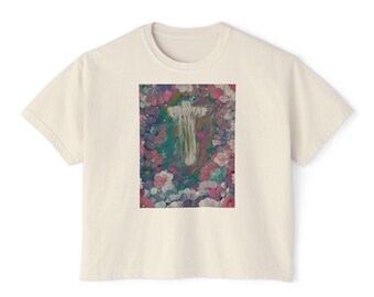 May include: A cream-coloured short-sleeve t-shirt featuring a rectangular graphic print on the chest. The print displays a white cross centred amidst a vibrant, painterly background of blended pink, purple, green, and blue floral-like shapes.