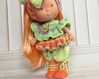 Op de afbeelding: Een handgemaakte pop met een gebreide outfit. De pop heeft lang oranje haar en draagt een kleurrijk ensemble van groen, oranje en paars. De outfit bestaat uit een hoed met een strik, een top, een korte broek en laarzen. De laarzen zijn oranje met witte strepen.