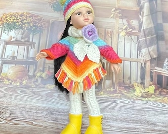 Può includere: Una bambola con lunghi capelli scuri indossa un berretto in maglia a righe arcobaleno con un grande pon-pon, un poncho colorato con motivo a chevron e frange, un fiore all'uncinetto bianco, leggings a trecce bianchi e stivali gialli.