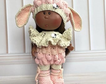 Op de afbeelding: Pop gekleed in een gebreid schaapskostuum, voornamelijk roze en crème. De outfit bestaat uit een hoed met lange oren, een top met ruches, een korte broek en laarzen. Kleine schapenmotieven zijn aanwezig op de hoed en top.