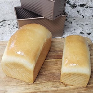 Può includere: Due pagnotte di pane appena sfornato sono su un tagliere di legno, accanto a tre teglie per pane color rame. Il pane ha una crosta dorata e una superficie morbida e lucida. Le teglie sono impilate e hanno un design a coste.