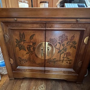May include: A wooden cabinet with two doors, featuring floral and branch carvings. The doors have brass hardware and a circular handle. The cabinet is a warm brown color and sits on a wooden floor.