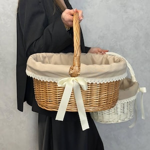 May include: Two wicker baskets. The larger basket has a woven handle and is lined with beige fabric and lace trim. A cream-colored bow adorns the front. A smaller, white basket with a similar design is held alongside the larger one.