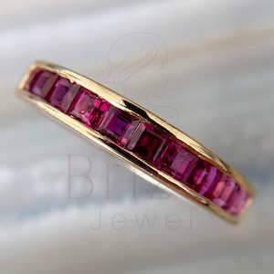 Puede incluir: Un anillo de banda de oro engastado con piedras preciosas rosas rectangulares y vibrantes. El diseño del anillo presenta un engaste en canal, mostrando las piedras preciosas en una fila continua. El anillo se muestra sobre una superficie suave y clara.
