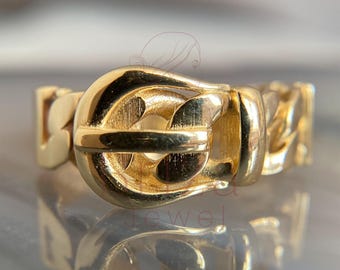 May include: A gold-colored ring with a buckle design. The ring features a chain-link band and a prominent buckle-shaped centerpiece. The polished metal reflects light, highlighting the intricate details of the jewelry.