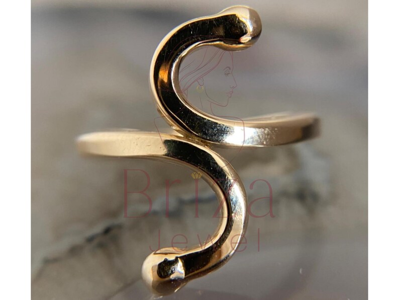May include: A gold-colored ring with a unique, abstract design. The ring features a curved, snake-like shape with rounded ends. The ring's design is smooth and polished, reflecting light. The ring is a piece of jewelry.