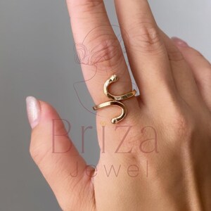 May include: A gold-colored ring in a snake design. The ring wraps around the finger with a curved, open design. The ring is on a hand with a light skin tone. The ring is a piece of jewelry.