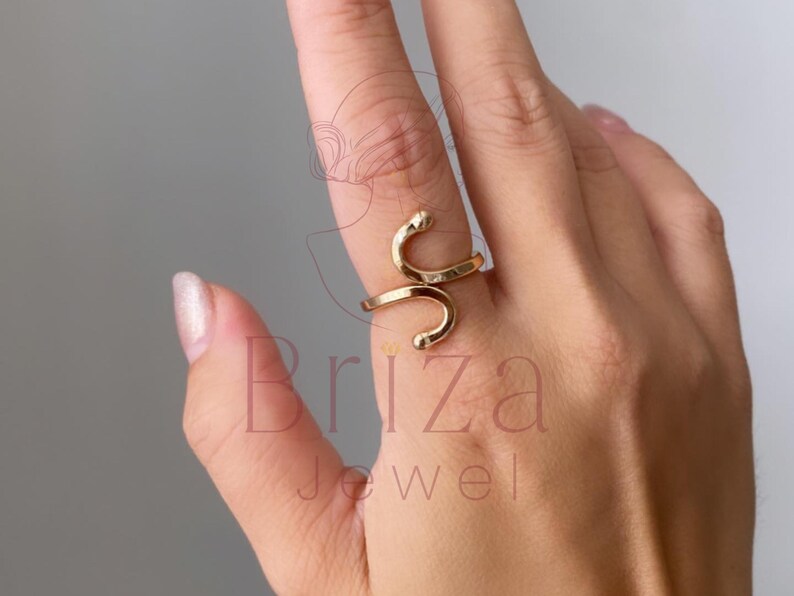 May include: A gold-colored ring in a snake design. The ring wraps around the finger with a curved, open design. The ring is on a hand with a light skin tone. The ring is a piece of jewelry.