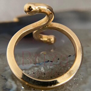 May include: A gold-colored ring with a snake design. The ring features a circular band with a snake head and body curving above the band. The ring is set against a neutral background.