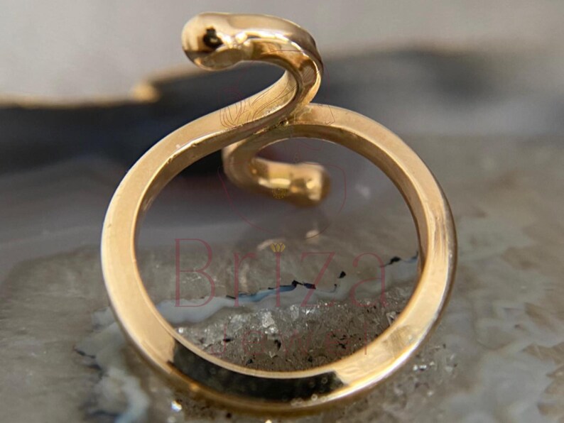 May include: A gold-colored ring with a snake design. The ring features a circular band with a snake head and body curving above the band. The ring is set against a neutral background.
