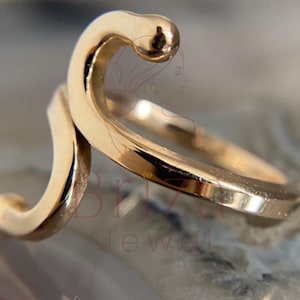 May include: A gold-colored ring with a unique, flowing design. The ring features a curved, wave-like shape with a small, rounded detail at the top. The ring's surface reflects light, highlighting its smooth texture. Jewelry.