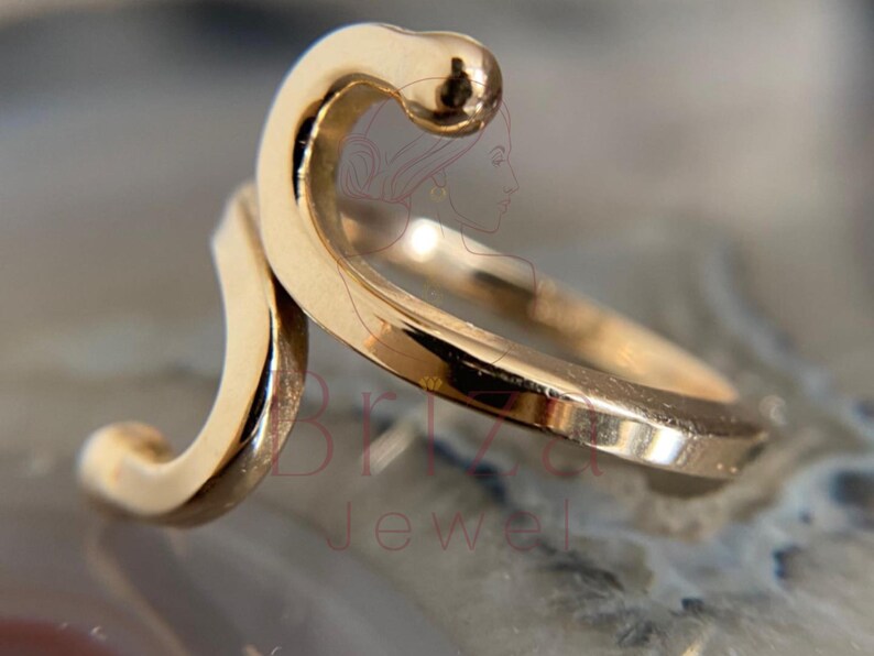 May include: A gold-colored ring with a unique, flowing design. The ring features a curved, wave-like shape with a small, rounded detail at the top. The ring's surface reflects light, highlighting its smooth texture. Jewelry.