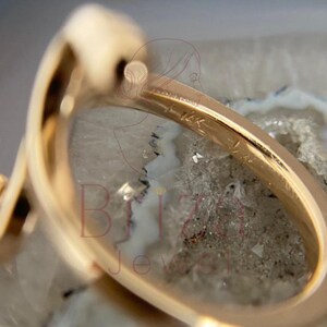 May include: Close-up of a gold ring with a unique, curved design. The ring is marked with "14K" and features a smooth, polished surface. The ring is set against a neutral, textured background, highlighting its elegant form.