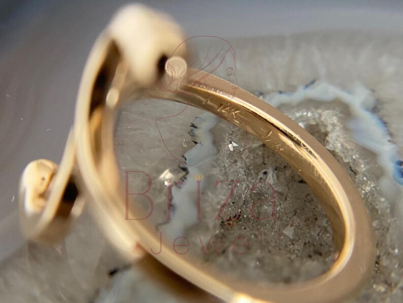 May include: Close-up of a gold ring with a unique, curved design. The ring is marked with "14K" and features a smooth, polished surface. The ring is set against a neutral, textured background, highlighting its elegant form.