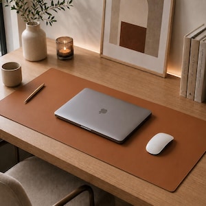 Op de afbeelding: Een rechthoekige bureauonderlegger van bruin leer op een houten oppervlak. De mat heeft een glad, effen oppervlak met afgeronde hoeken en biedt een vlakke werkruimte voor een zilveren laptop, een witte computermuis en een goudkleurige pen.