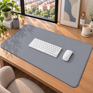 Peut inclure: Un grand tapis de bureau gris, lisse et rectangulaire, posé sur une surface en bois. Au centre du tapis se trouvent un clavier sans fil blanc et une souris d'ordinateur sans fil blanche. Le tapis présente des coins arrondis et une texture uniforme.