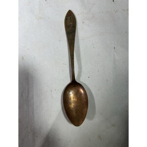 May include: A tarnished copper-toned metal spoon featuring an elongated handle with a pointed tip. The handle displays a small, engraved graphic of a ram or goat near the top, followed by vertical text running down the stem.