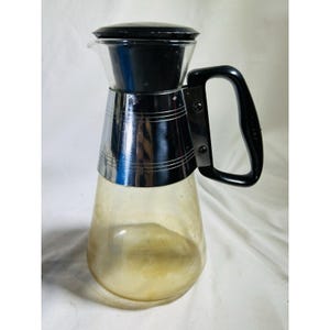 May include: A vintage glass pitcher with a black lid and handle. The pitcher has a chrome band around the middle and a clear glass body with a slight yellow tint. The handle is black and attached to the chrome band.