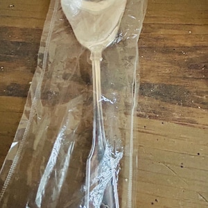 May include: A silver serving spoon in a clear plastic wrapper. The spoon has a wide, oval bowl and a long, slender handle. The handle features a decorative design at the base.