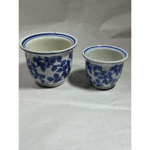 May include: Two white ceramic bowls of different sizes, each featuring a blue vine and leaf pattern around the exterior and blue circular bands around the inner and outer rims. The bowls have a smooth, rounded shape and a white interior finish.