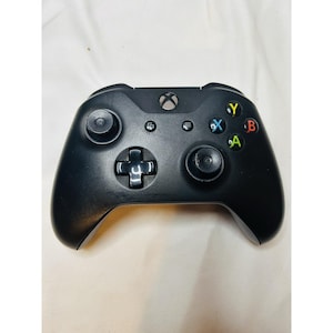 Puede incluir: Mando Xbox negro con botones de colores (verde A, rojo B, azul X, amarillo Y). Cuenta con dos palancas analógicas, una cruceta y botones de función. El logotipo de Xbox es visible en la parte superior.