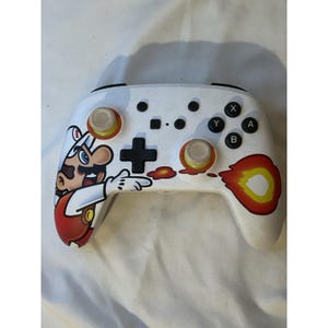 May include: White video game controller with a custom Mario design. The controller features a cartoon image of Mario on the left side, with orange and yellow accents. The buttons are black, and the joysticks are orange.