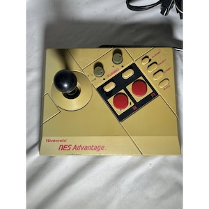 May include: A tan Nintendo NES Advantage controller with a black joystick, red A and B buttons, and a Turbo button. The controller has a rectangular shape with the text "NES Advantage" printed on the front.