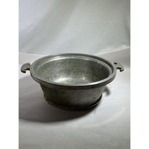 May include: A vintage metal bowl with a textured exterior and two small handles. The bowl has a wide rim and a slightly tapered shape. The metal has a dull, silver color, suggesting it may be made of aluminum or a similar alloy.
