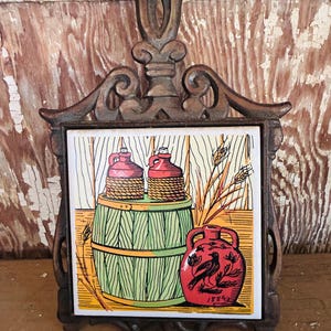 May include: A decorative trivet with a cast iron frame and a ceramic tile insert. The tile features a colorful illustration of two jugs atop a green barrel, a red jug with a bird design, and wheat stalks. The trivet is designed to protect surfaces from heat.