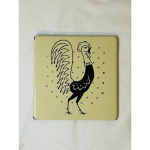 May include: Square ceramic trivet with a rooster design. The rooster is black with a detailed tail and a comb. The background is a light yellow with small black crosses scattered across the surface. The trivet has a glossy finish.