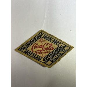 May include: A diamond-shaped fabric patch with a tan background and black border. It features the red Coca-Cola logo, the text 75th ANNIVERSARY 1902-1977, BOTTLED REFRESHMENT, and DALLAS, TX. The embroidery is stitched in red and black thread.