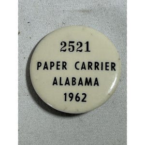 May include: A round, off-white button with black text. The text reads "2521 PAPER CARRIER ALABAMA 1962". The button is likely a commemorative item or pin.