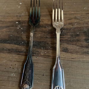 May include: Two antique silver forks with ornate shell designs on the handles. One fork is tarnished, displaying a darker, aged appearance, while the other is polished and shiny. The forks are displayed on a rustic wooden surface.
