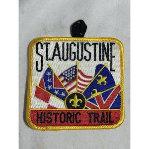 May include: Embroidered patch with the text "ST AUGUSTINE HISTORIC TRAIL". The patch features flags, including the American flag, and a gold fleur-de-lis emblem. The border is yellow.