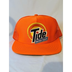 May include: Orange trucker hat with a mesh back and a curved brim. The front features a Tide logo in a yellow and orange circle, with the words "Tide Racing Team" below.